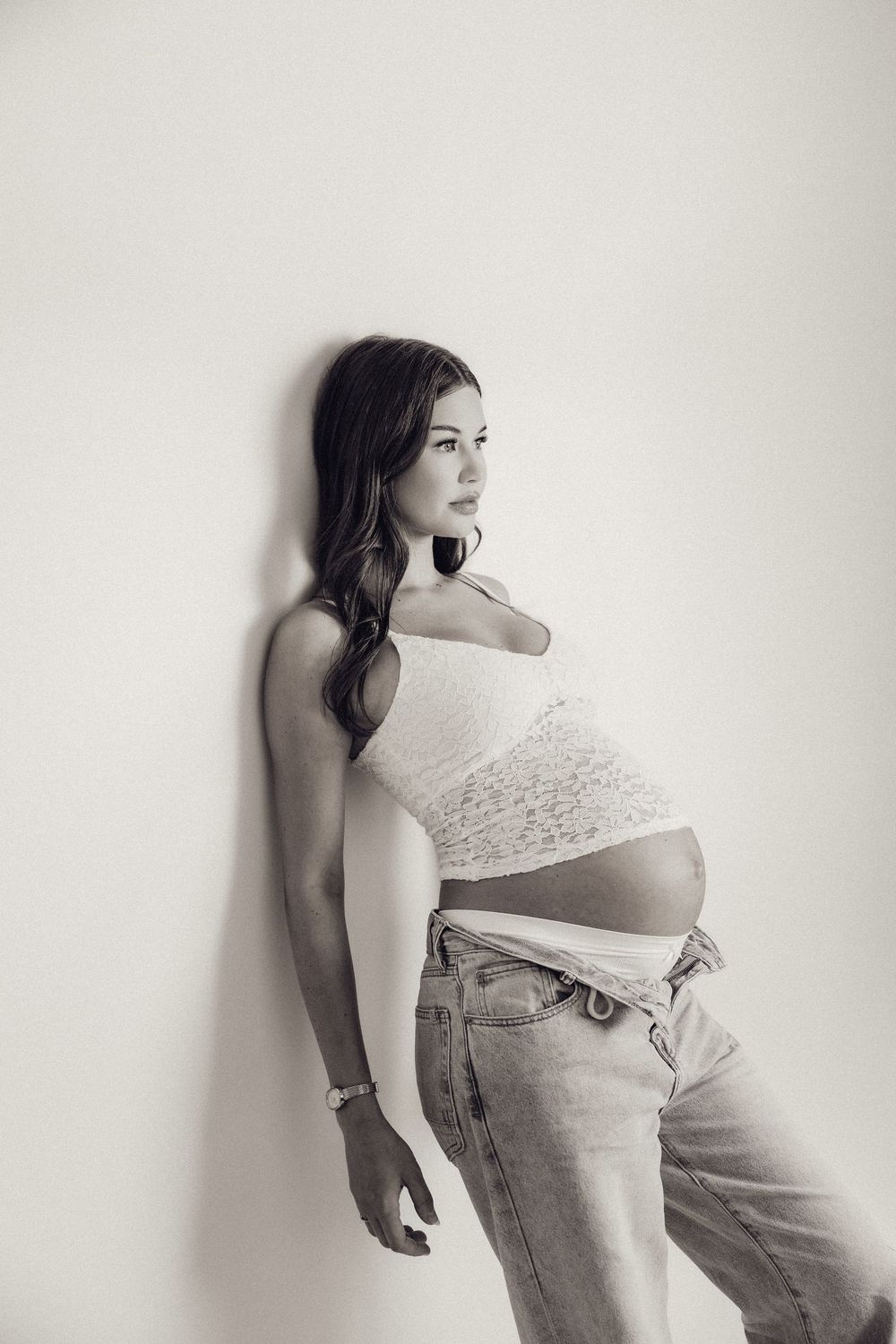 Black and white photo of a pregnant woman in jeans and lace crop top leaning against a white wall.