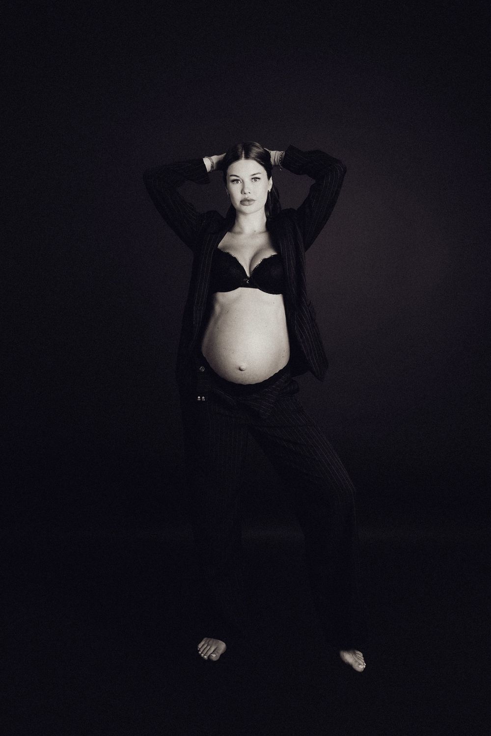 Pregnant woman standing in black bra and trousers, hands in hair, showing bare bump in moody black and white studio photo.