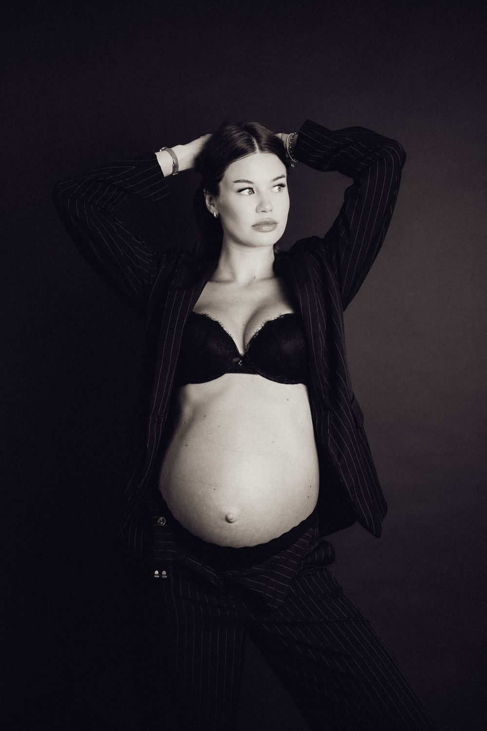 Black and white photo of pregnant woman in open pinstripe blazer and black bra showing baby bump.