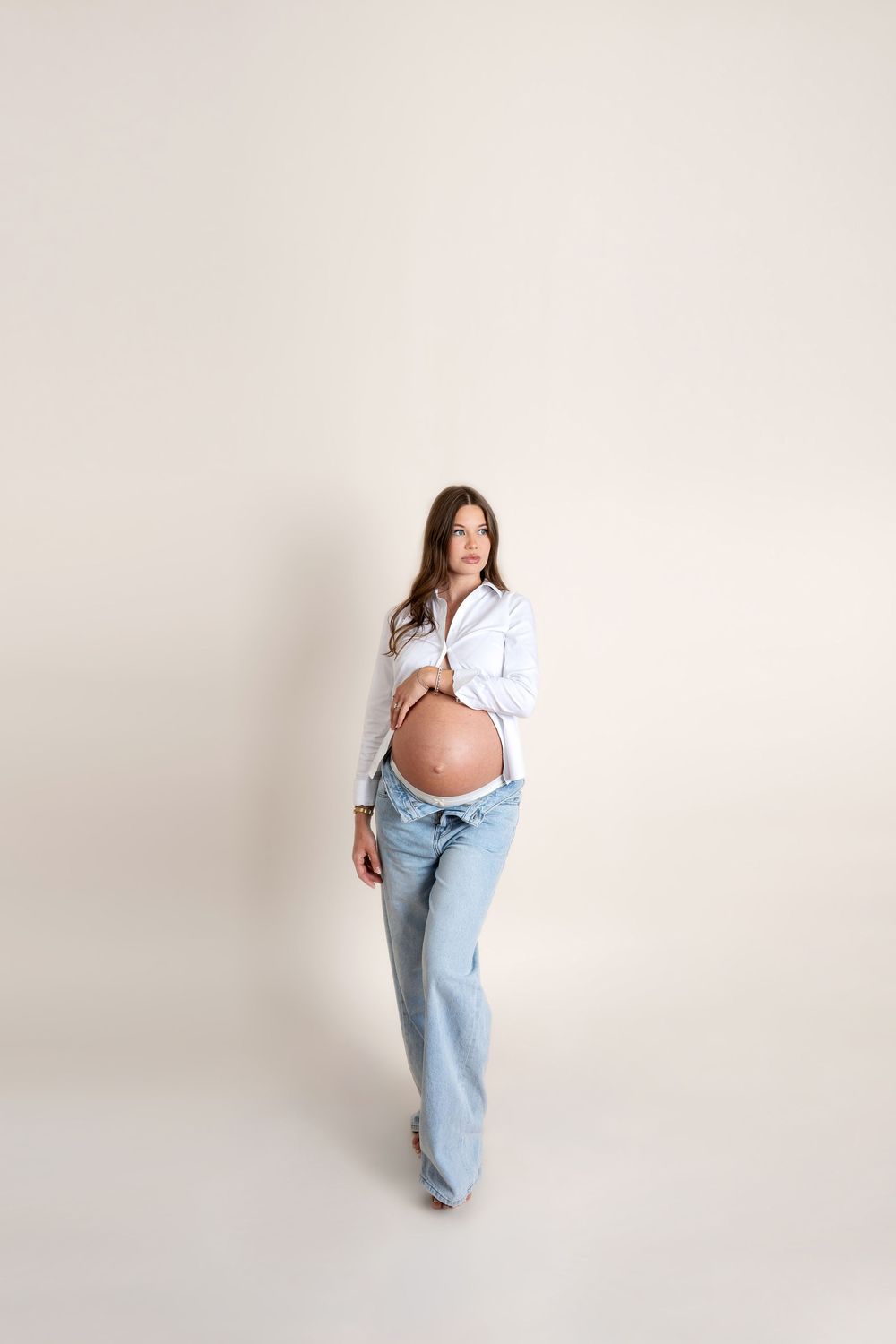 Pregnant woman in an open white shirt and low-rise jeans stands showing her bare baby bump on a light background.