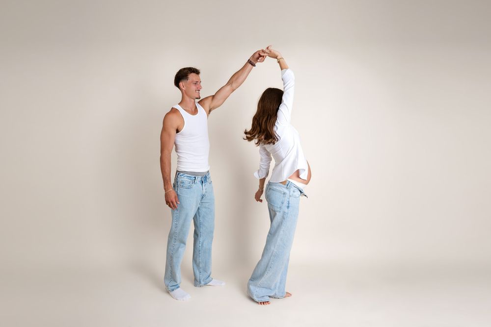A couple dances together in matching white tops and light blue jeans against a neutral background.