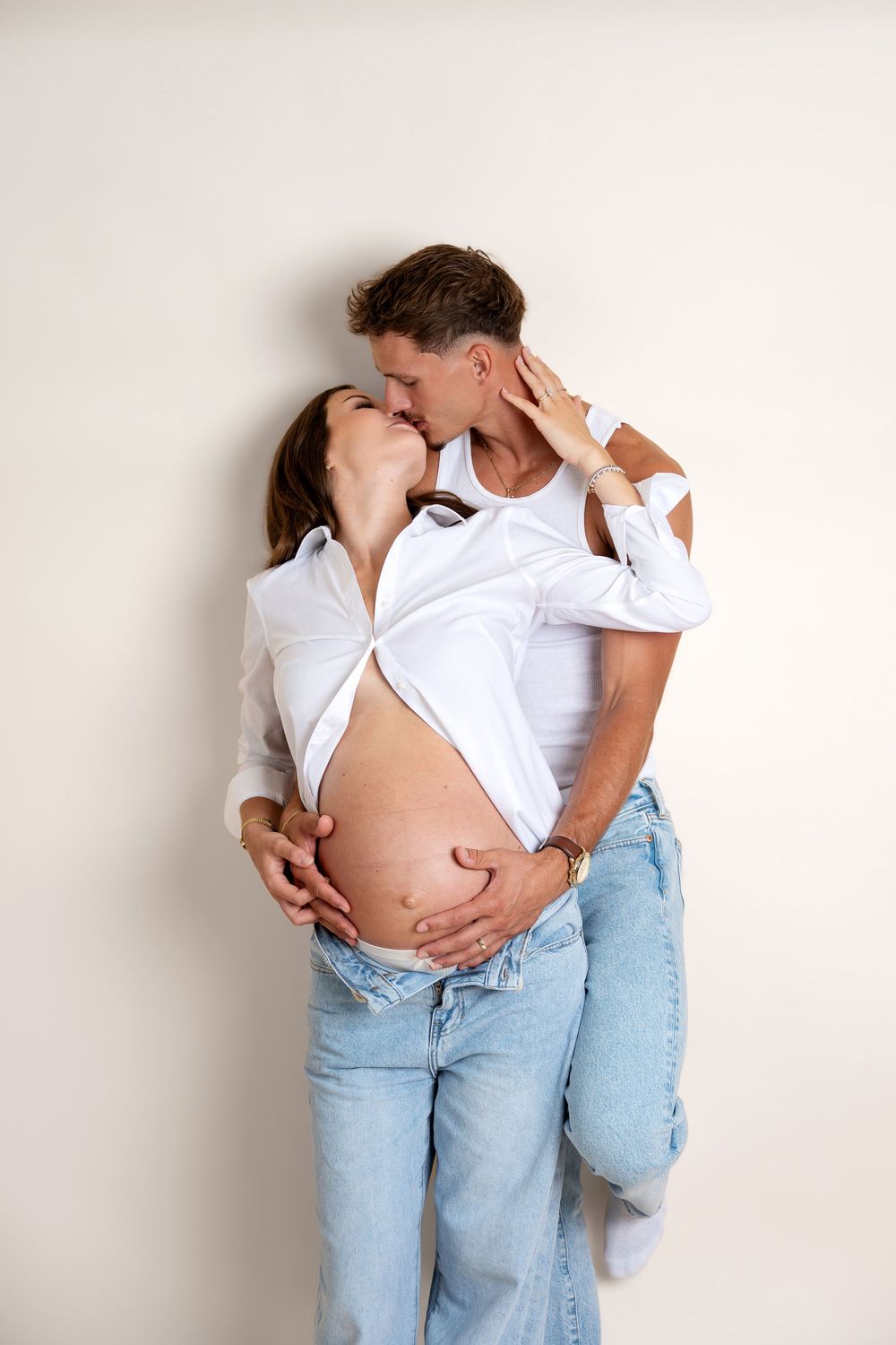 Expecting couple kissing tenderly, man cradling pregnant belly, dressed in white tops and blue jeans.