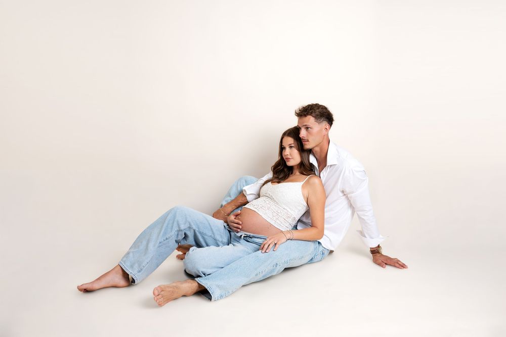 Couple sitting on floor, pregnant woman leaning on man who embraces her baby bump.