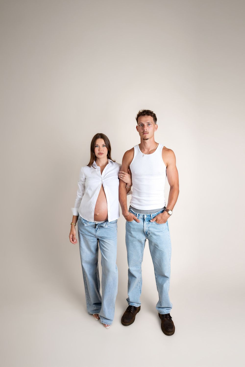 Pregnant woman in open white shirt and jeans standing with partner in white tank top against light background.