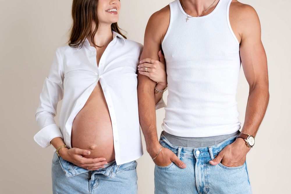 Cropped maternity photo of couple in matching white shirts and jeans, pregnant woman cradling her baby bump.