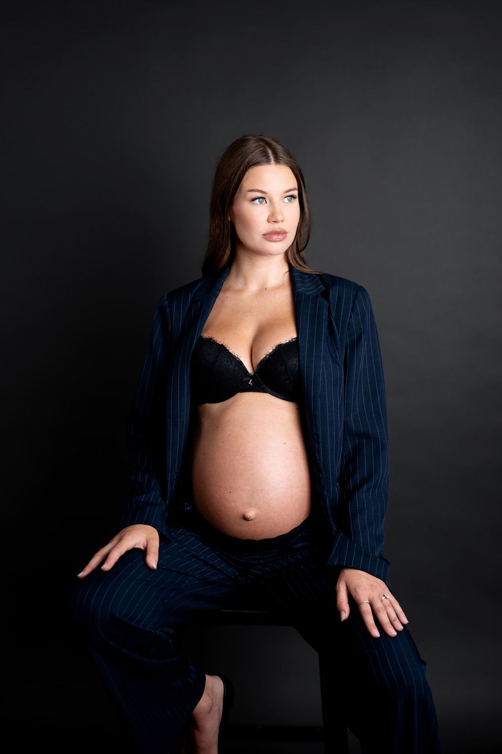 Pregnant woman in navy pinstripe suit and black lace bra sitting and showing bare baby bump.