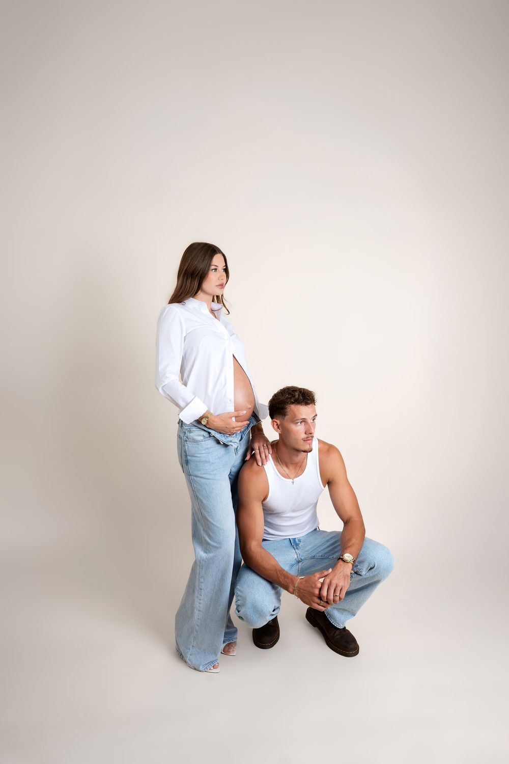 Pregnant woman in white shirt and jeans posing with man in white tank and jeans in studio.