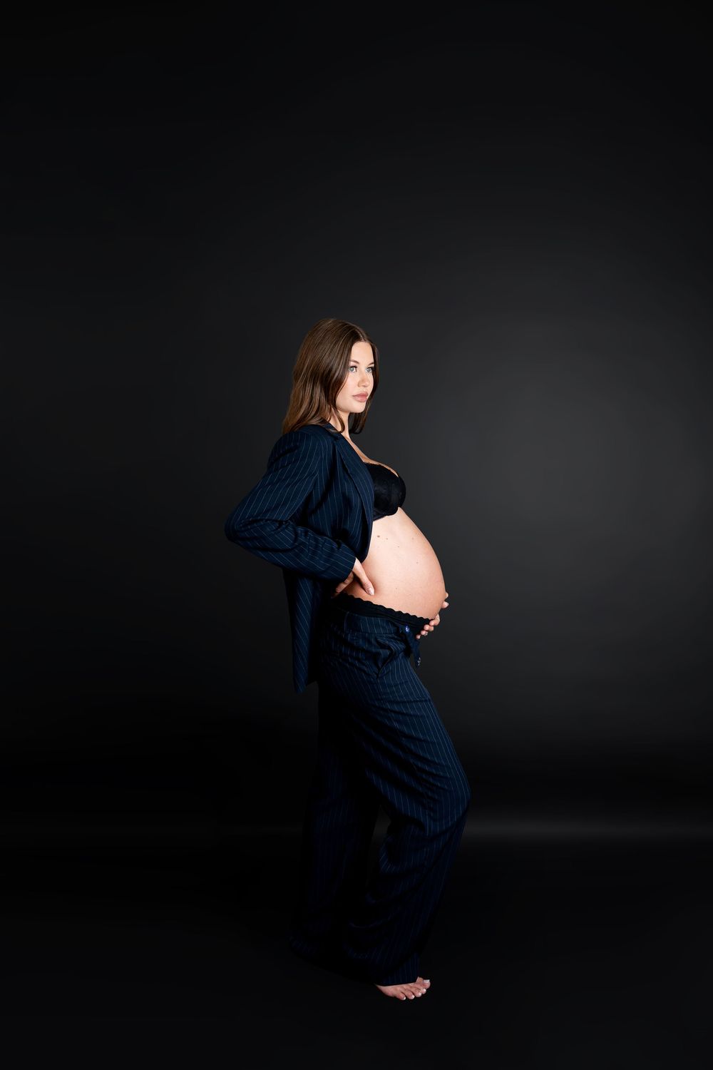 Pregnant woman in navy blue suit posing sideways showing baby bump against dark background.