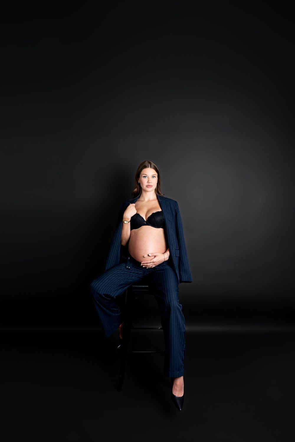 A pregnant woman in a navy suit and black bra sits on a stool posing in a dark studio.