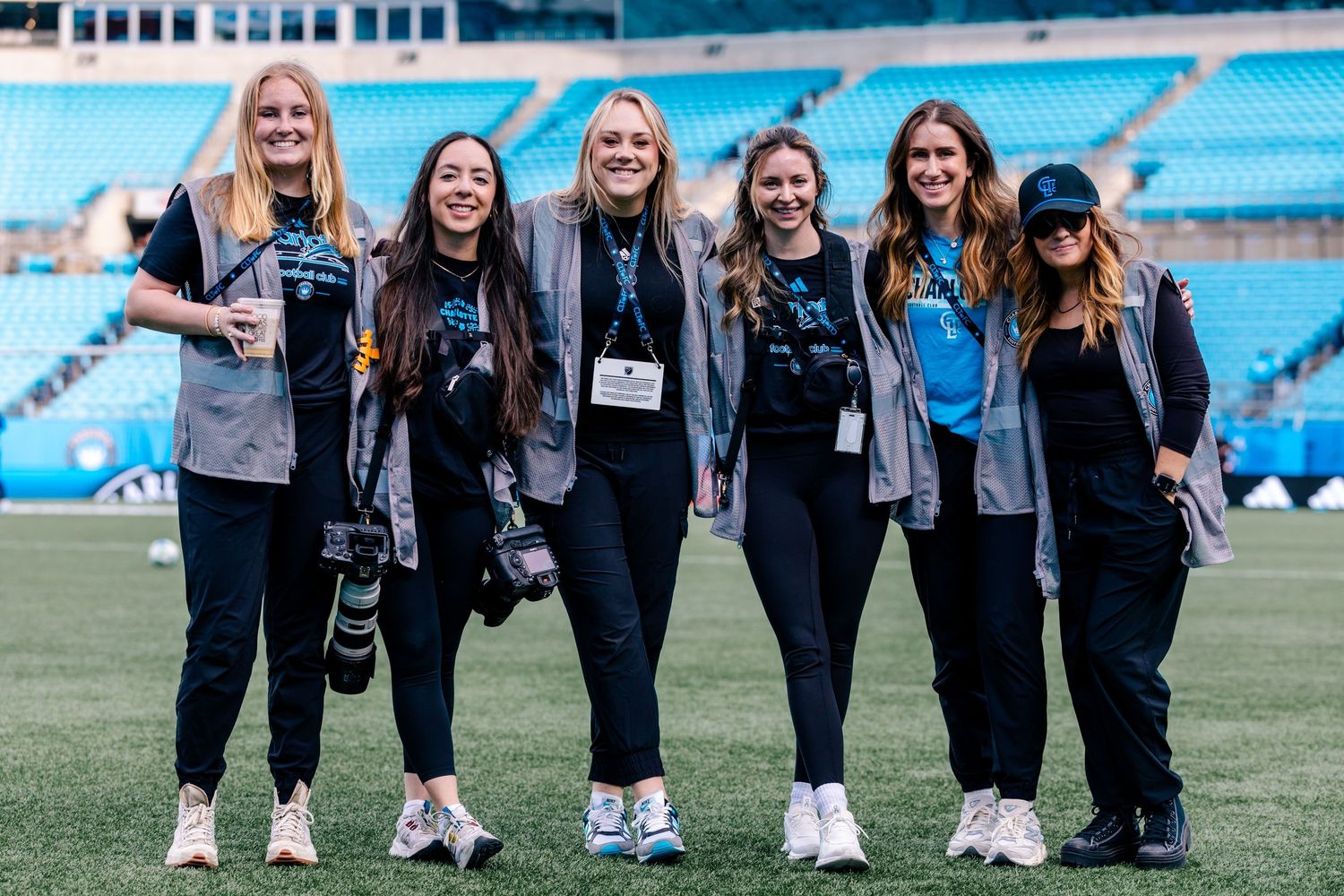 Six women wearing lanyards pose together smiling on a soccer field inside a stadium.