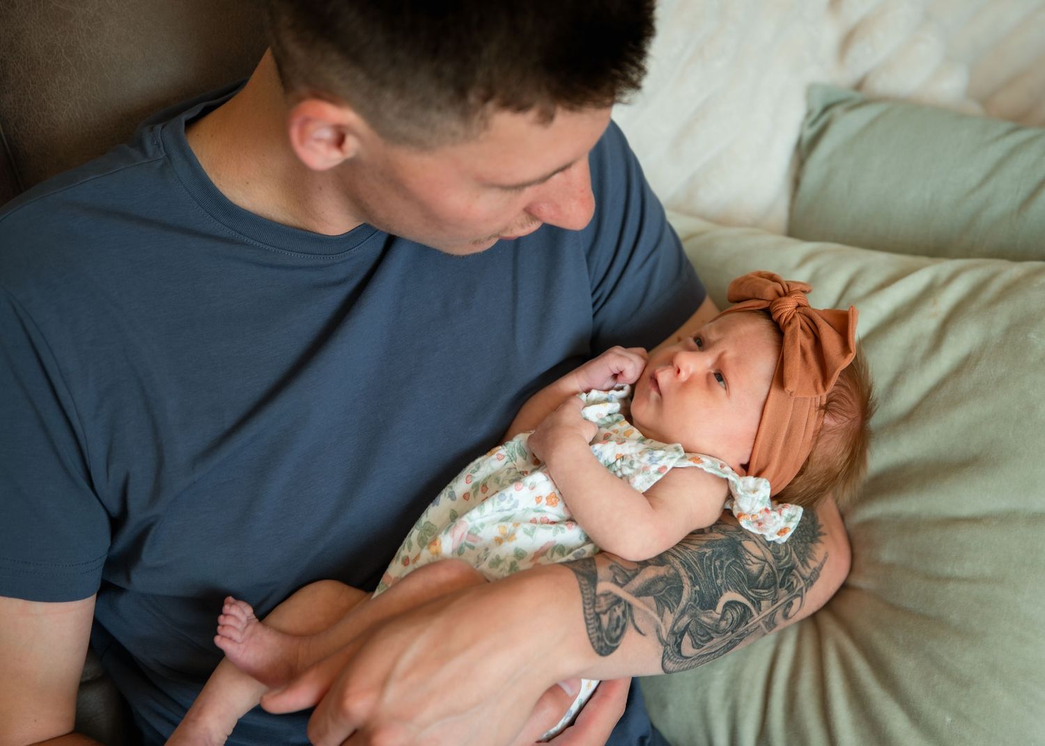 A tattooed father tenderly holds his newborn baby with a red bow headband while reclining on a green pillow.