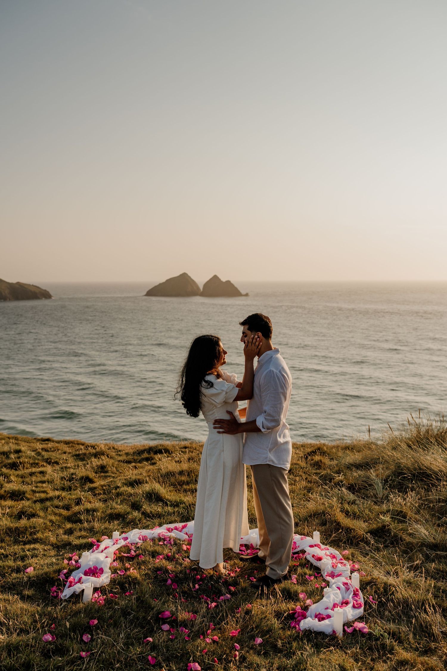 Couple embracing at sunset on coastal cliffside with flower petal heart arrangement and ocean rocks in background.