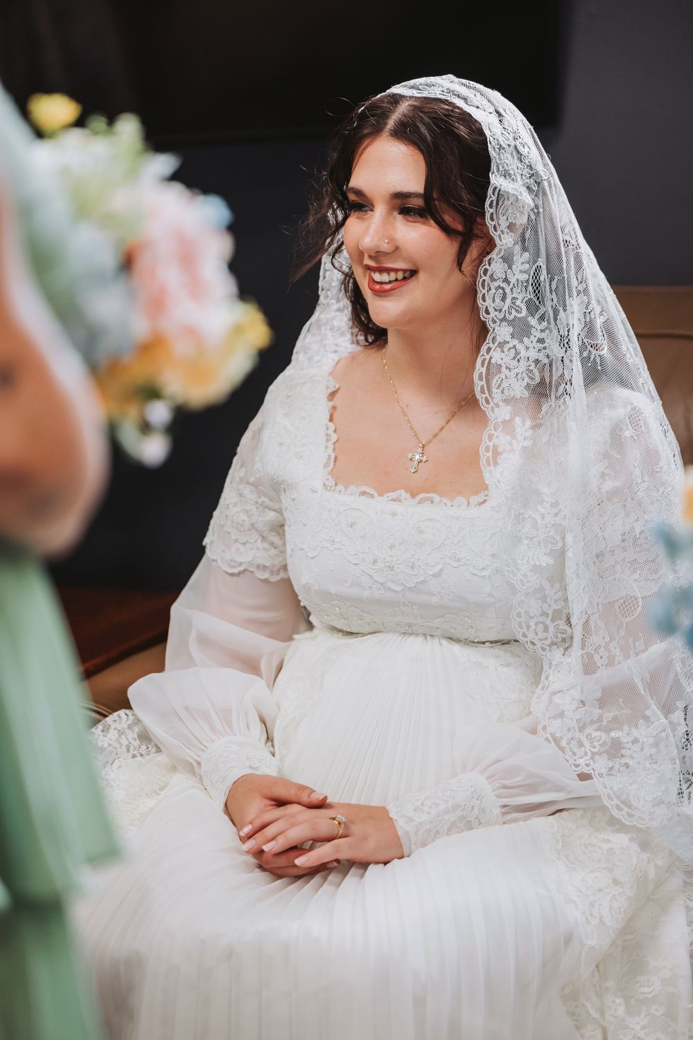 Smiling bride in white lace wedding dress and veil seated, wearing cross necklace, with colorful bouquet nearby.