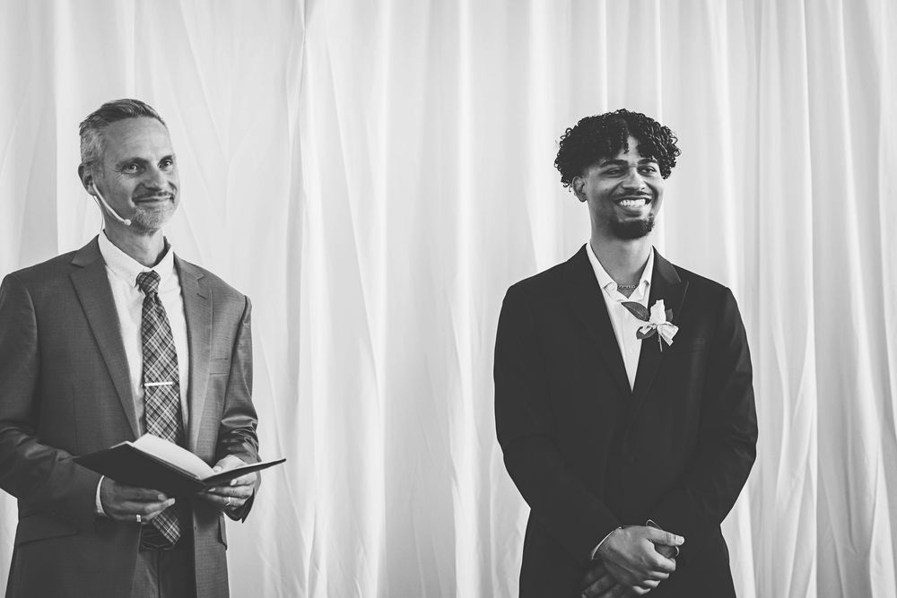Two men smiling during a wedding ceremony, one holding a book, in elegant suits, black and white photo.