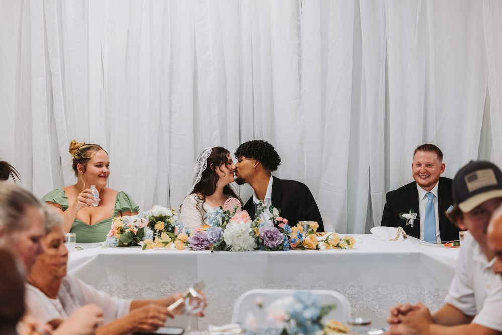 Bride and groom share a kiss at wedding reception head table while guests smile and celebrate around them.