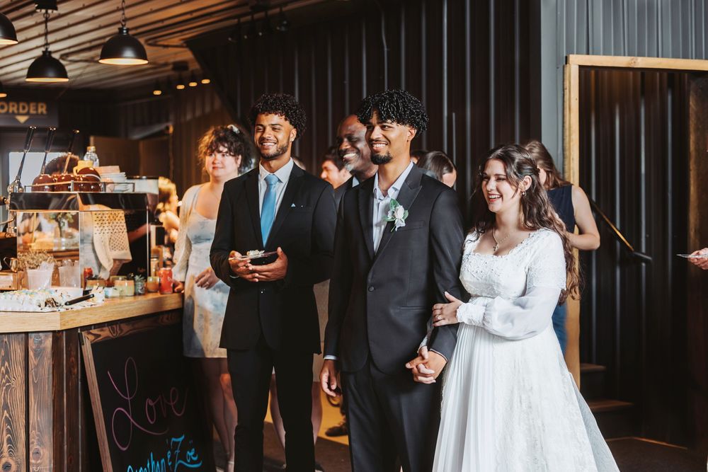 Bride in white gown and groom in dark suit smile during wedding reception at rustic venue with guests nearby.