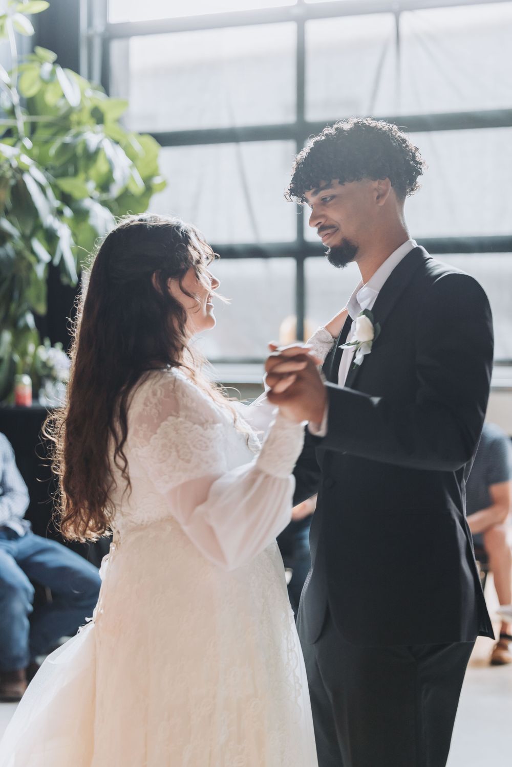 Bride and groom share their first dance at a wedding reception, holding hands and smiling in a sunlit venue.