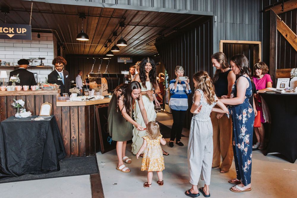 Women and children mingle at an indoor event space with a pickup counter and rustic wooden decor.