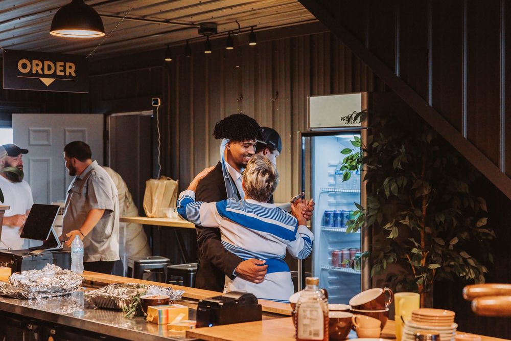 Two people share a warm hug inside a rustic bar or cafe with an ORDER sign and refrigerator in background.