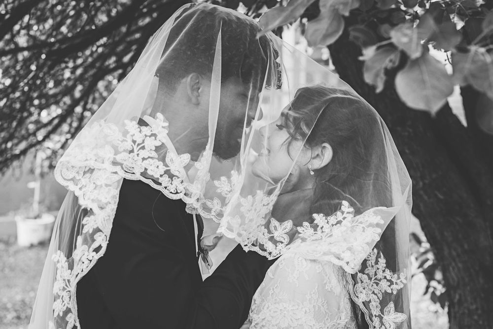 Bride and groom share romantic moment under lace wedding veil in black and white photo.