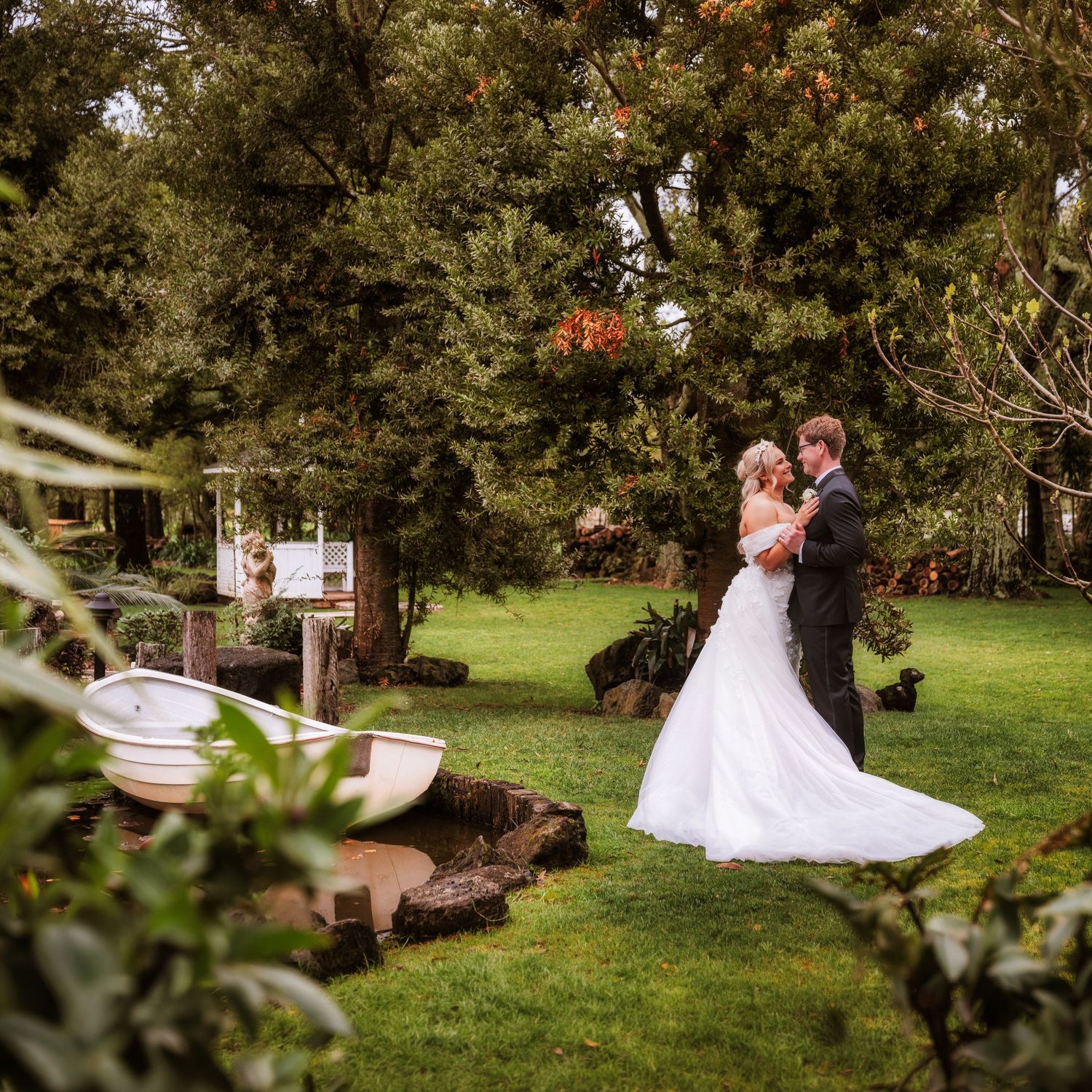 Bride and groom share a romantic embrace in a lush garden setting with a small pond and boat nearby.