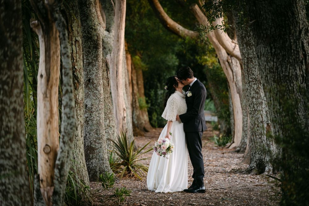 A bride and groom share a romantic kiss among tall trees on a forest path on their wedding day.