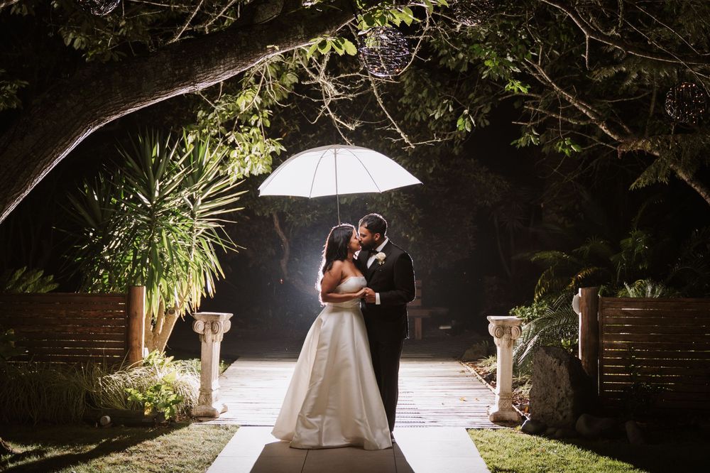 Bride and groom share a romantic kiss under a white umbrella on a candlelit garden path at night.