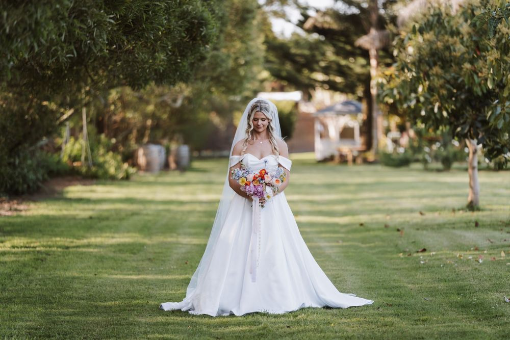 Bride in white off-shoulder gown and veil holds colorful bouquet on green lawn surrounded by trees.