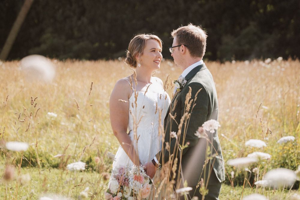 Couple sharing a romantic moment in a sunlit wildflower meadow, bride in white dress and groom in suit smiling at each other.