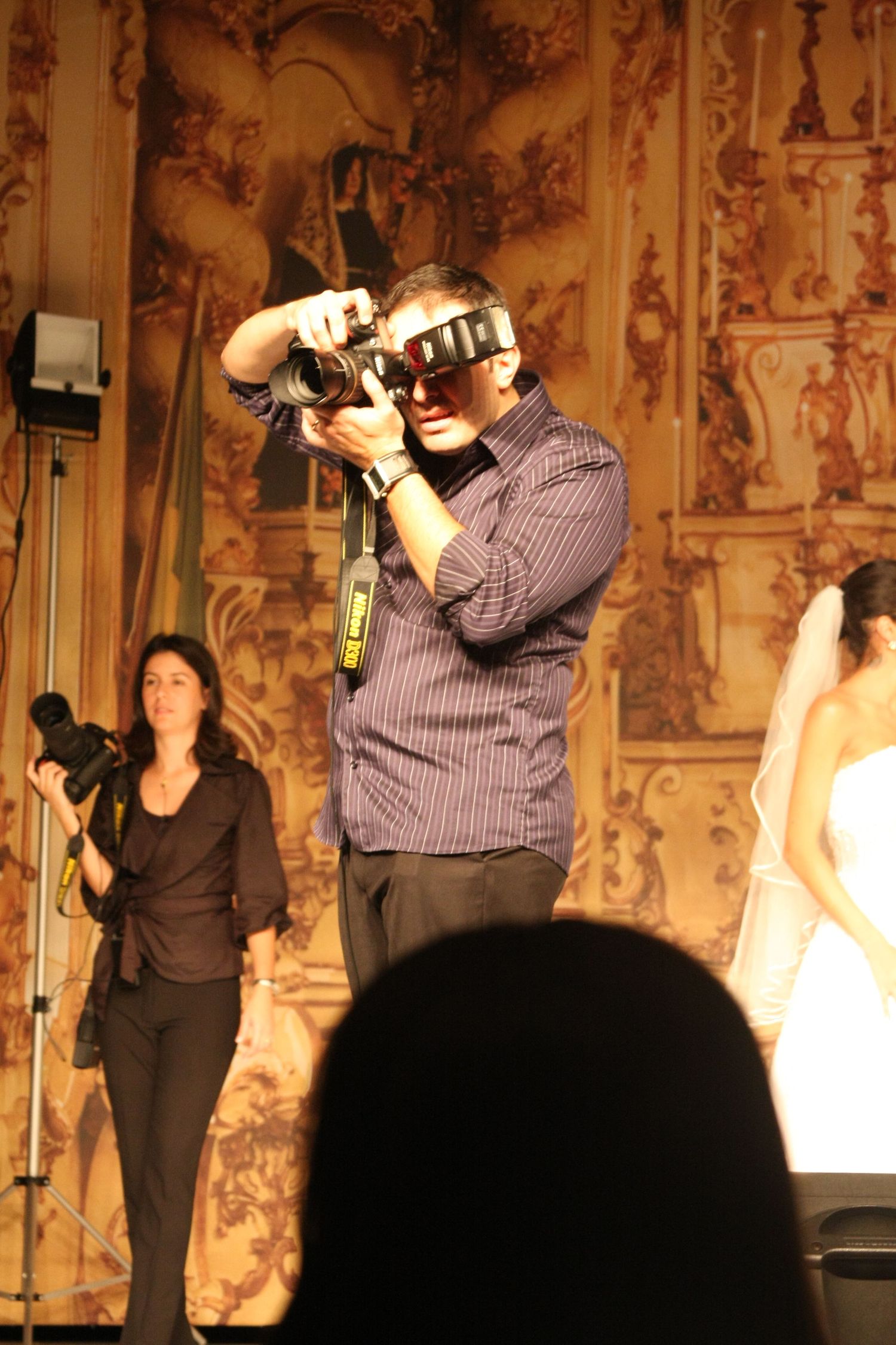 Photographer using a camera at a wedding event with ornate baroque backdrop and assistant holding camera behind him.