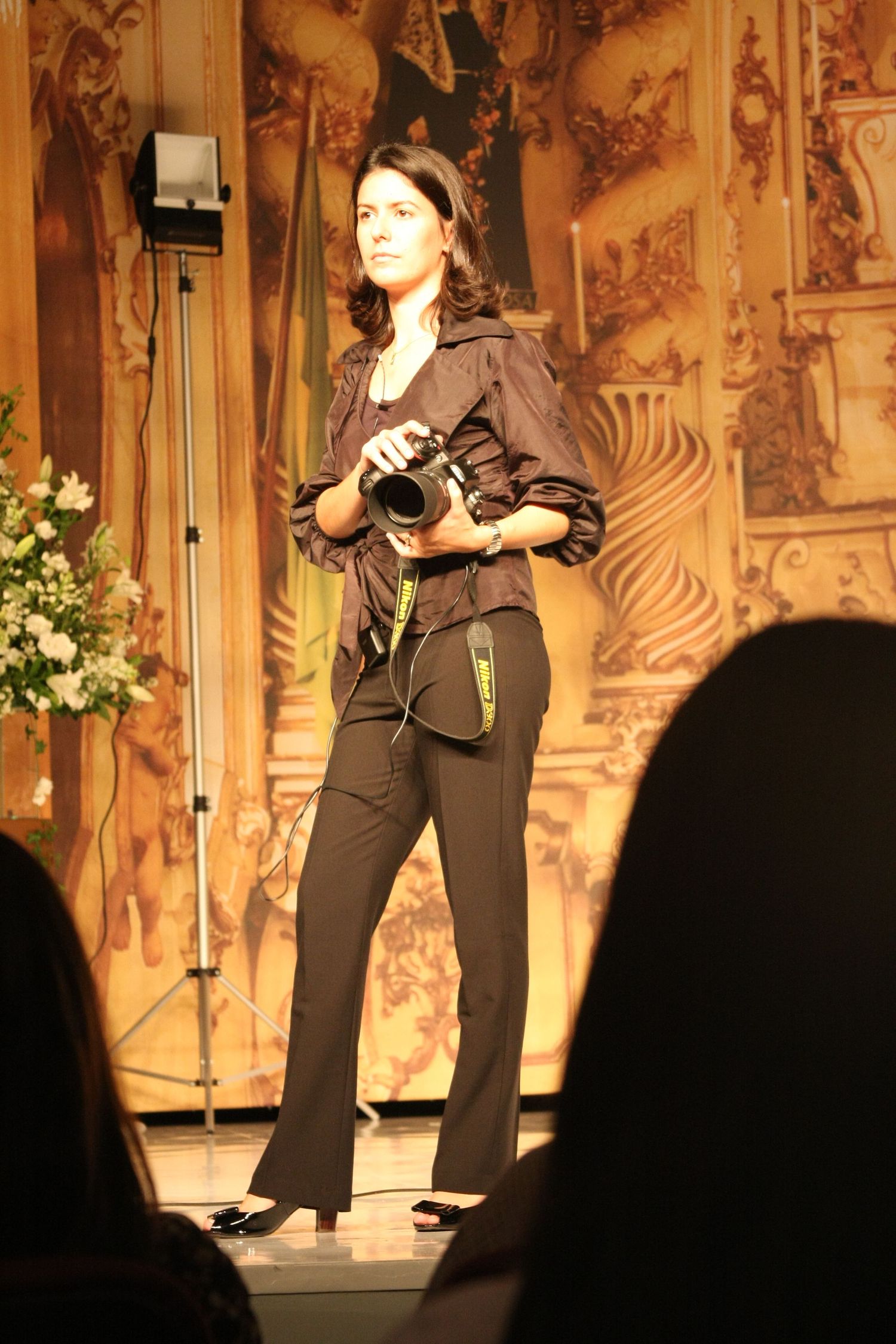 A woman photographer stands on stage holding a camera, wearing dark clothing against an ornate golden backdrop.