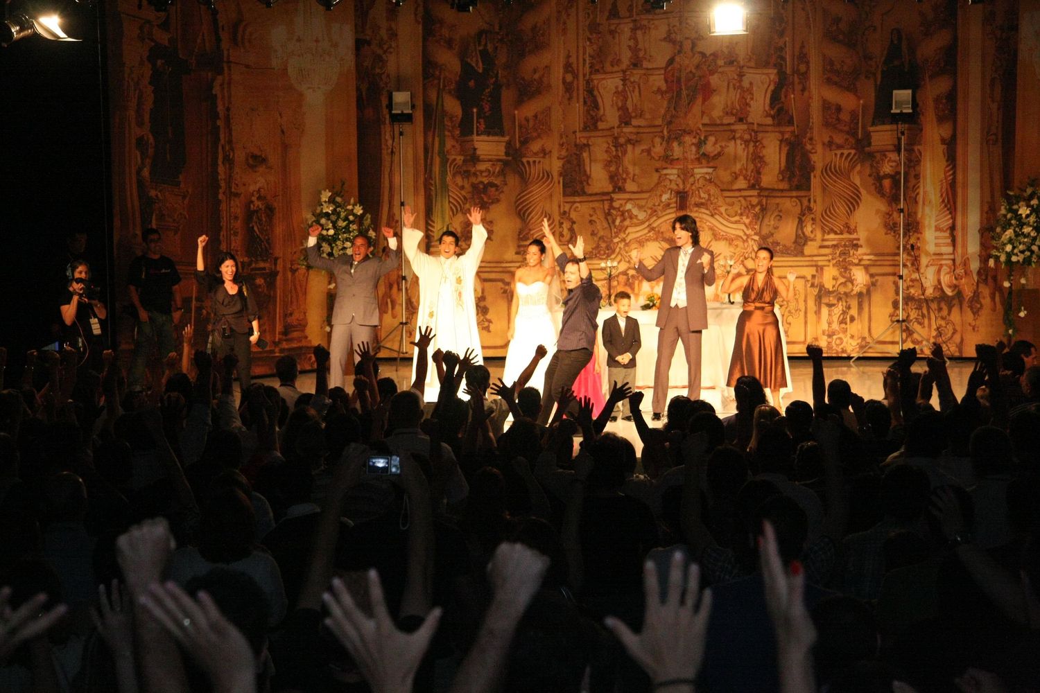 Performers take a bow on stage before an enthusiastic crowd with hands raised at a ornate theater venue.