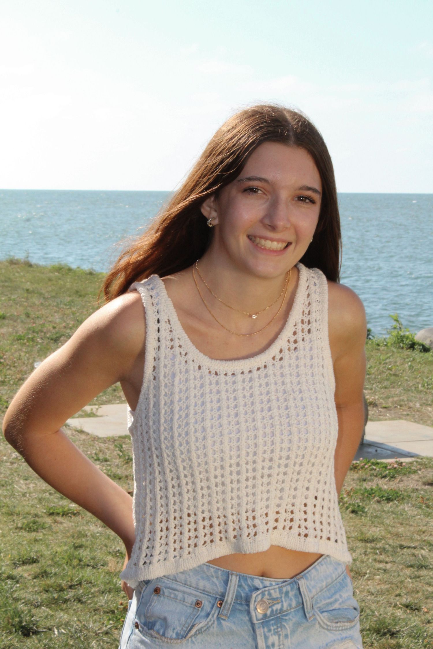 Smiling teenage girl in a white crochet top and jeans posing outdoors near the ocean.