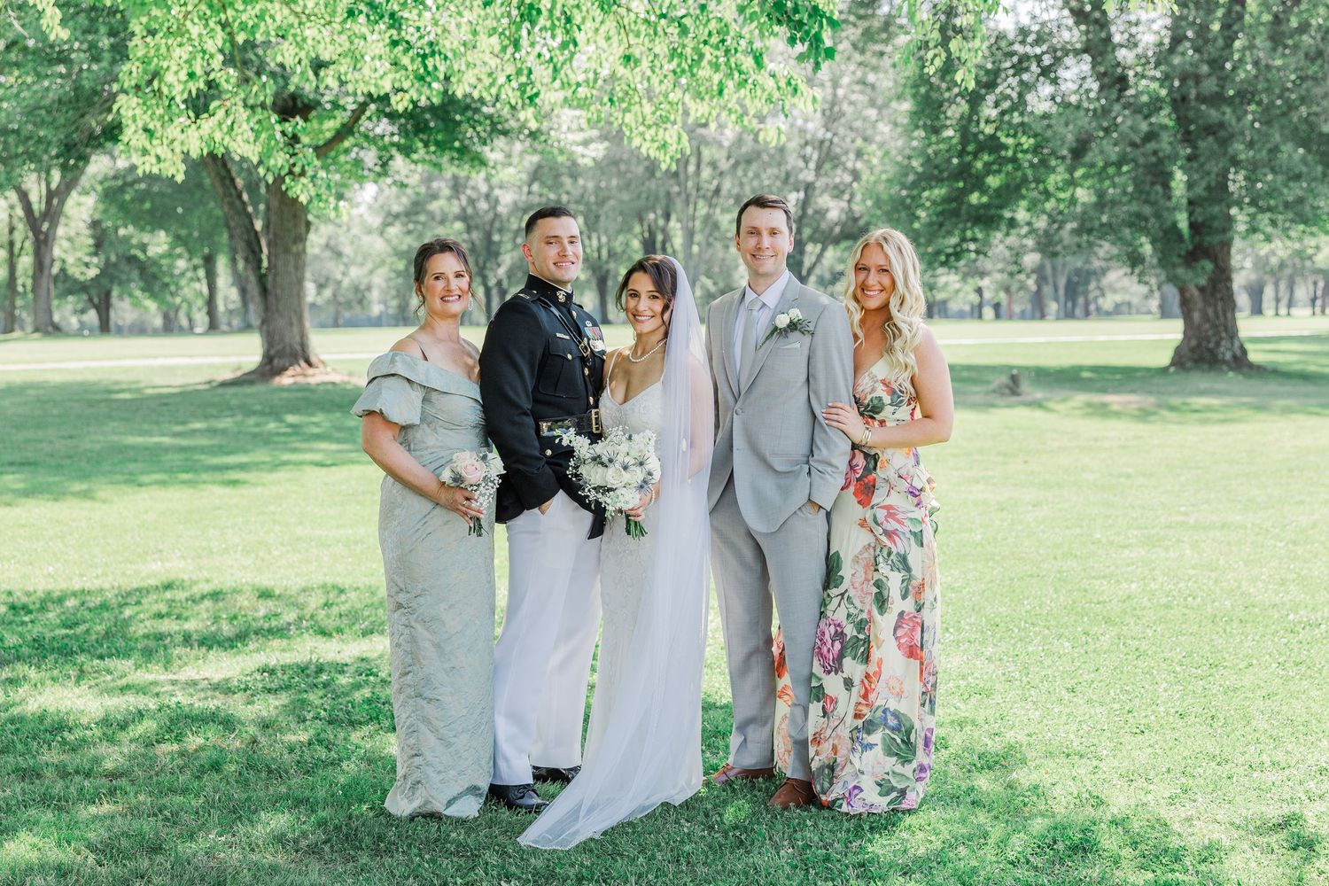 Wedding party poses outdoors in a lush green park, featuring bride in white, groom in gray suit, and guests in elegant attire.