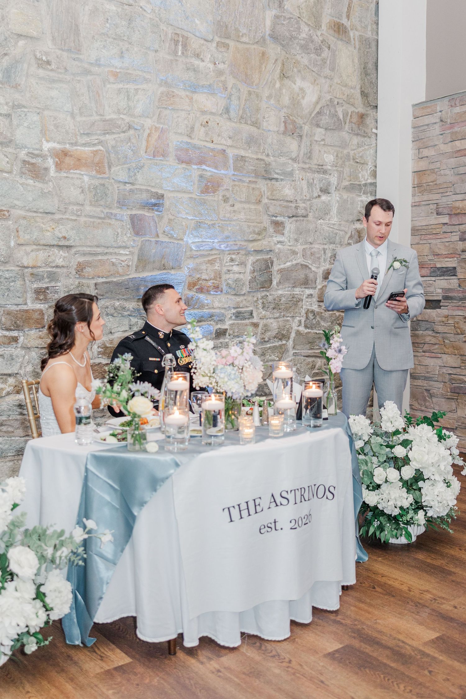 A couple sits at a sweetheart table decorated with florals and candles while a groomsman delivers a wedding toast.