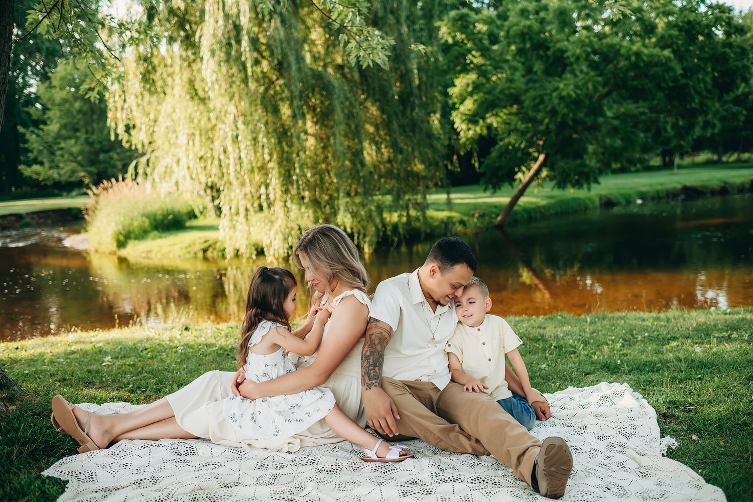 Family of four sitting on a blanket by a pond in a lush green park, sharing a tender moment together.