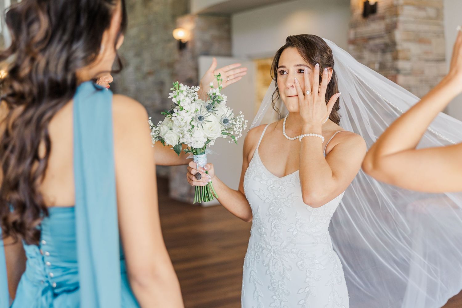 Bride in white gown adjusting veil while bridesmaid in blue dress holds white floral bouquet indoors.