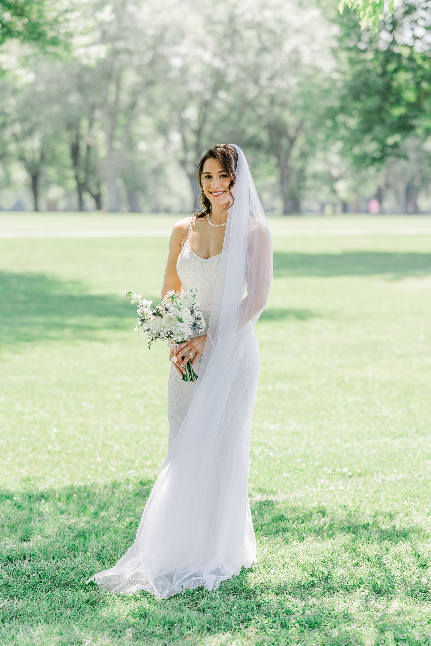 Bride in white lace gown and veil holds floral bouquet, smiling in a sunlit green park.