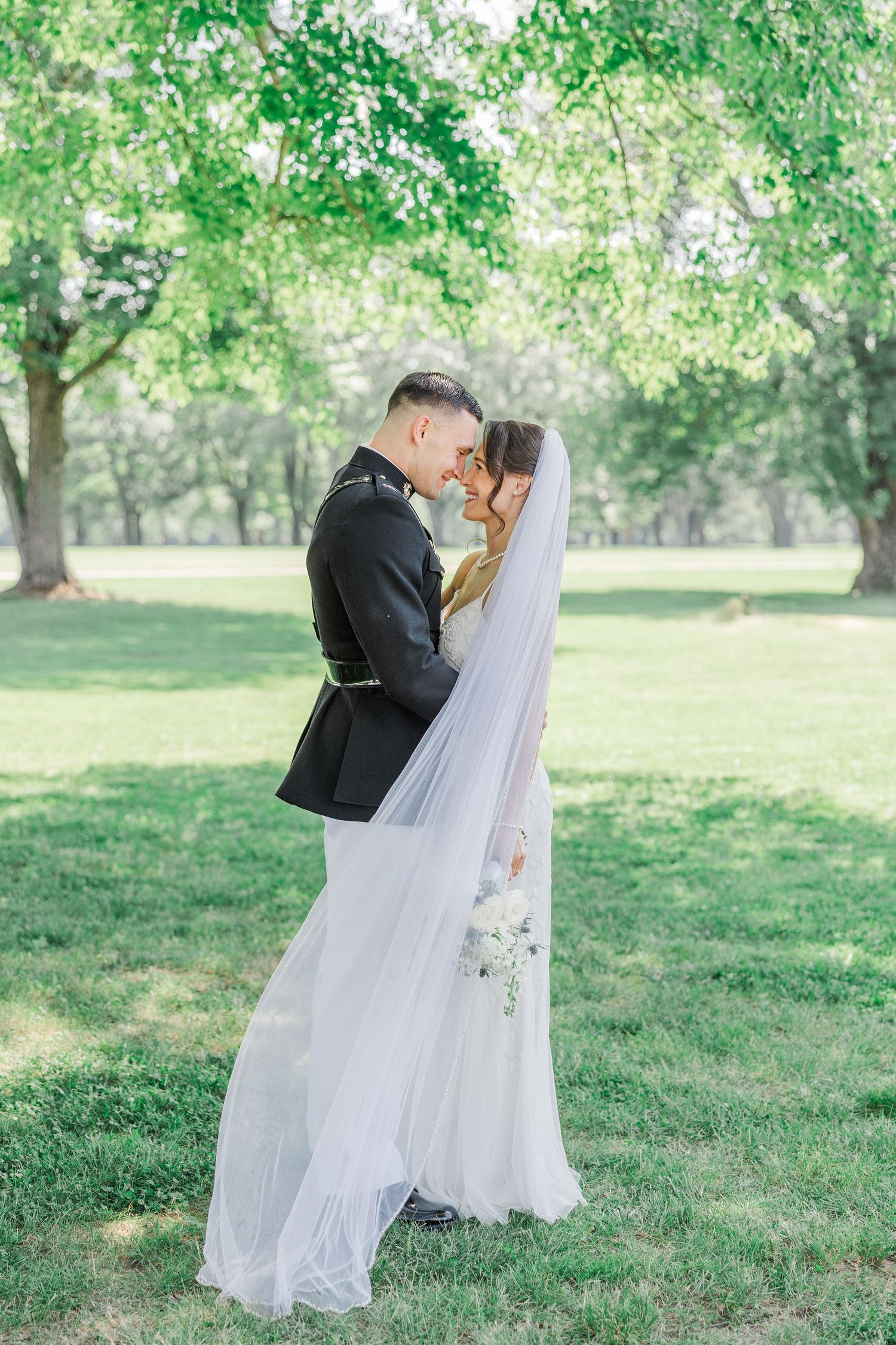 A bride and groom share a romantic moment in a lush green park, with her white veil flowing gracefully around them.