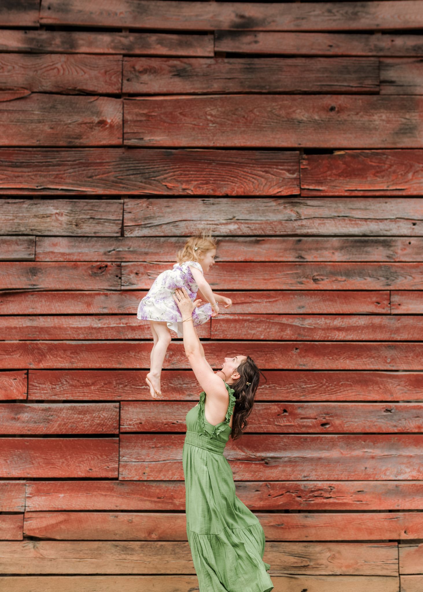A woman in a green dress joyfully lifts a toddler girl in the air against a rustic red barn wall.