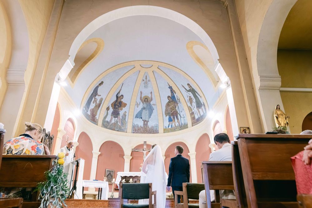 Couple stands at church altar beneath ornate dome fresco depicting saints, surrounded by wooden pews in elegant chapel.