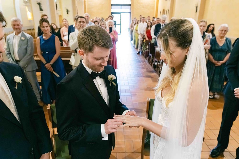 Groom placing wedding ring on bride's finger at church altar ceremony with guests watching in background.