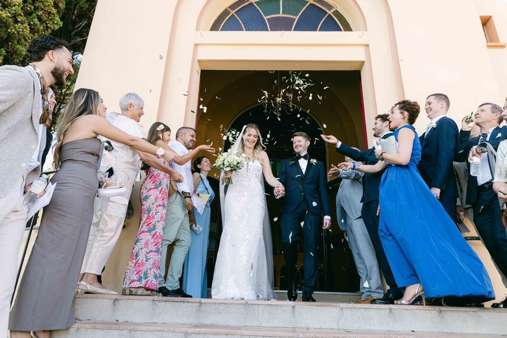 Newlywed couple exits church greeted by guests tossing flower petals in a joyful outdoor wedding ceremony.