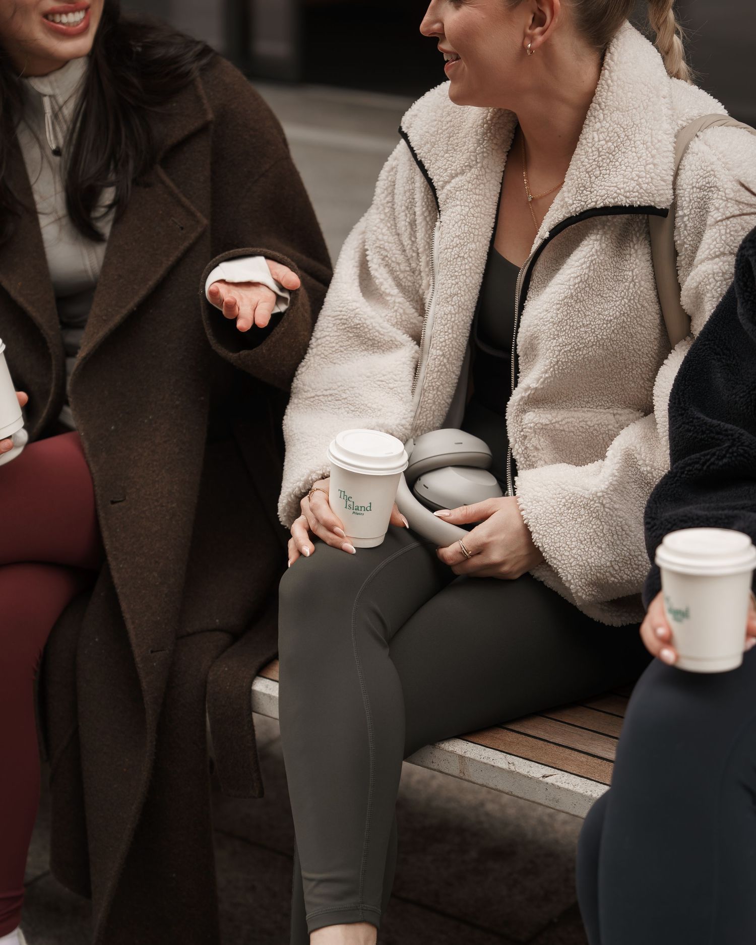 Women sitting on a bench outdoors, laughing and holding coffee cups, wearing cozy winter coats.