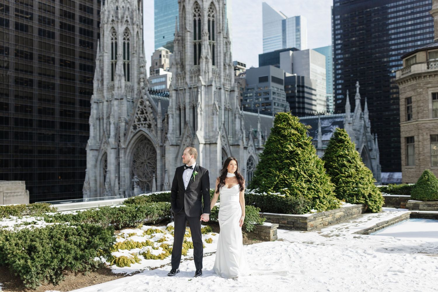 Bride and groom hold hands in snowy NYC plaza with St. Patrick's Cathedral and skyscrapers in background.