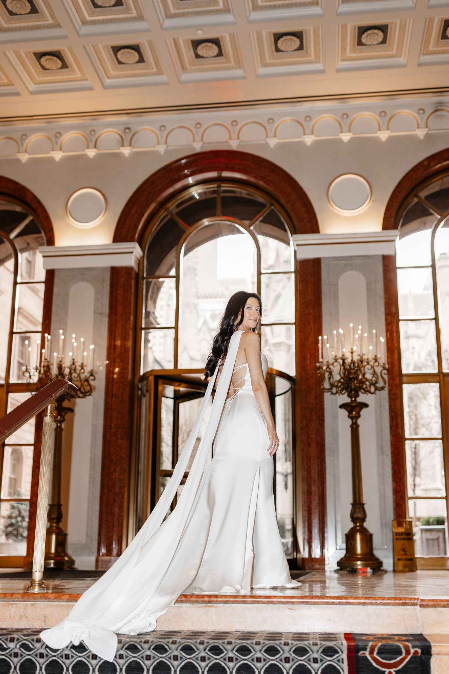 Elegant bride in a white gown with long veil stands in a grand ornate ballroom with arched windows and candelabras.