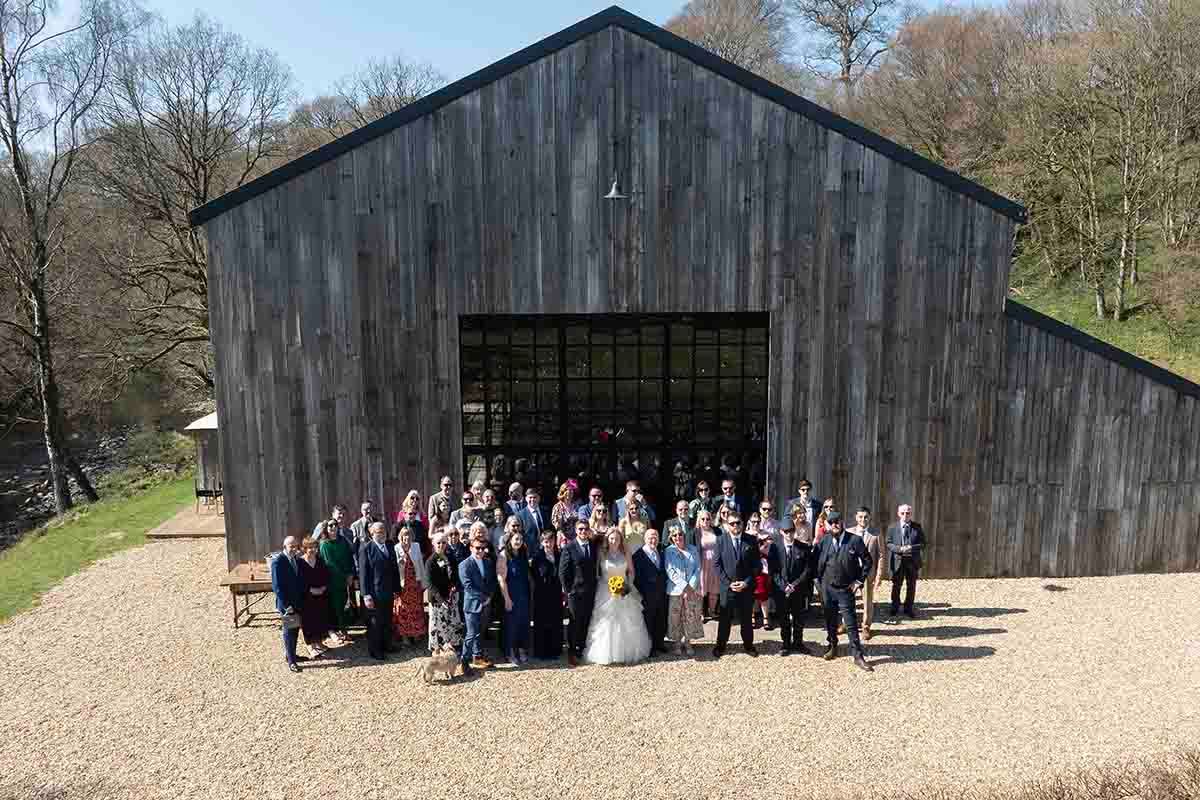 Large wedding group photo outside a rustic wooden barn venue on a sunny day with trees in the background.