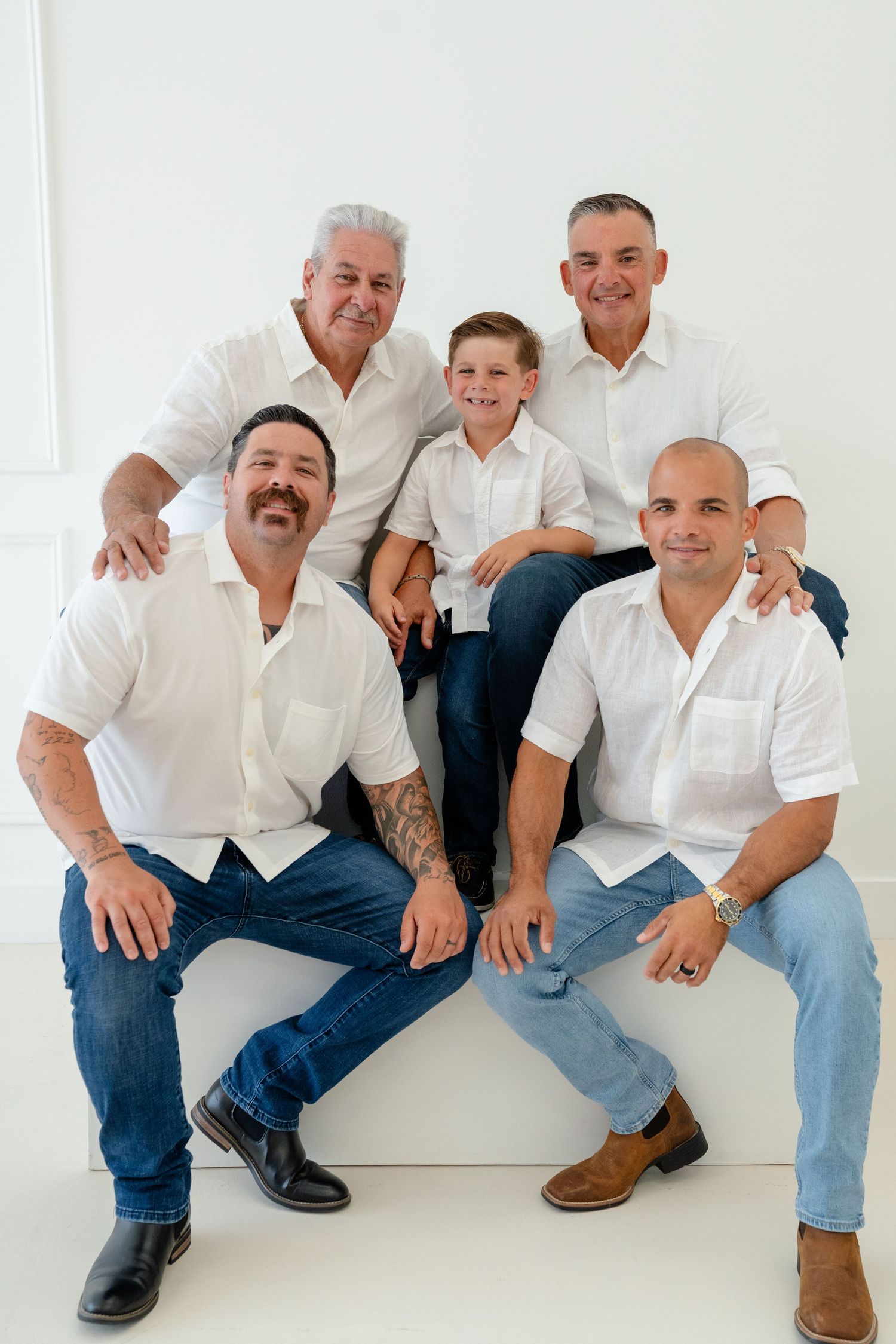Multi-generational family portrait of five males in white shirts and jeans posing together in a bright white studio setting.