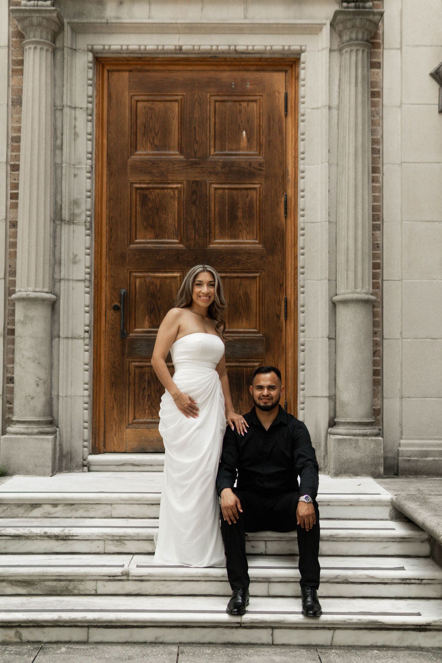 Bride in white strapless gown and groom in black suit posing on stone steps by wooden door.