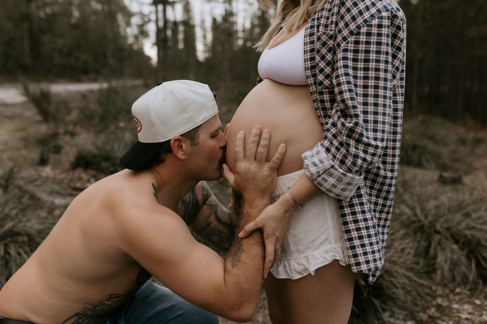 Man kissing pregnant womans baby bump outdoors in forest setting.